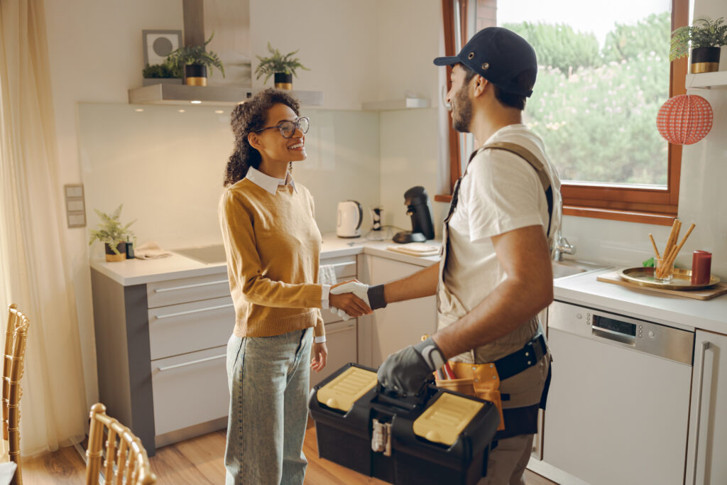 professional repairman in uniform shaking hands with woman while standing at home kitchen
